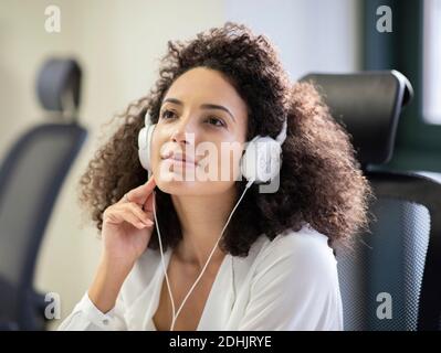 Jeune femme hispanique attentionnée, blouse blanche et casque regarder loin tout en travaillant à table avec ordinateur dans moderne lieu de travail Banque D'Images