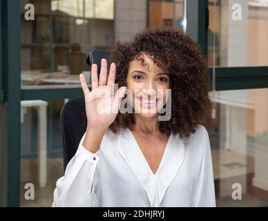Jeune femme d'affaires ethnique avec une coiffure afro assis au bureau tout en regardant l'appareil photo en agitant la main Banque D'Images