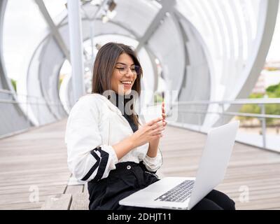 Vue latérale d'une blogueuse ethnique millénaire positive et moderne un café à emporter et un ordinateur portable assis sur un pont fermé au-dessus de la rivière Banque D'Images