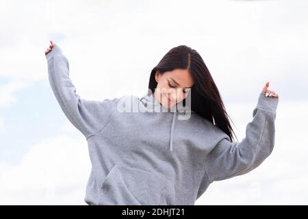 Faible angle de la jeune femme heureuse dans gris hoodie ayant amusez-vous et sautez avec les bras levés contre le ciel nuageux Banque D'Images