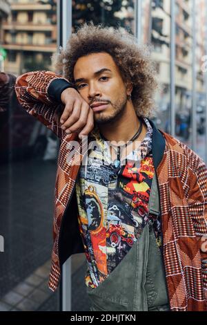 Beau jeune homme afro-américain barbu aux cheveux bouclés dans une tenue élégante et colorée, regardez l'appareil photo tout en vous appuyant contre paroi en verre de Banque D'Images