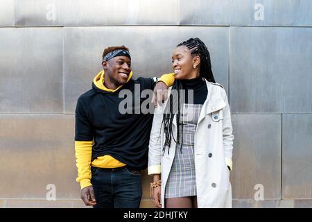Jeune couple afro-américain heureux avec des tenues tendance mur en métal se regardant l'un contre l'autre tout en se tenant contre contemporain bâtiment urbain Banque D'Images