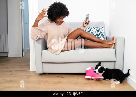 Vue latérale d'une jeune femme noire dans des vêtements décontractés assis sur un canapé avec un smartphone à portée de main et vue sur mignon petit chiot jouant avec botte à Banque D'Images