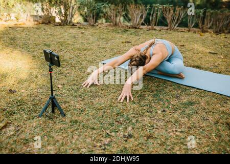 Une femme blogueuse enregistre des vidéos sur un smartphone pour les médias sociaux Pratiquer le yoga sur tapis à Baddha Konasana dans le parc d'été Banque D'Images