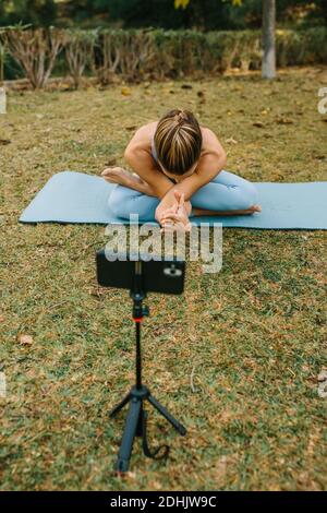 Une femme blogueuse enregistre des vidéos sur un smartphone pour les médias sociaux Pratiquer le yoga sur tapis dans Lotus pose dans le parc d'été Banque D'Images