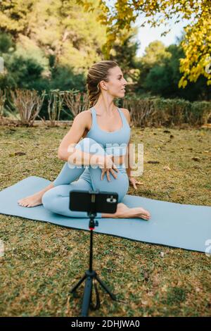 Flexible femme pratiquant le yoga et l'enregistrement vidéo sur smartphone pour Blog tout en étant assis à Ardha Matsyendrasana sur tapis dans le parc Banque D'Images