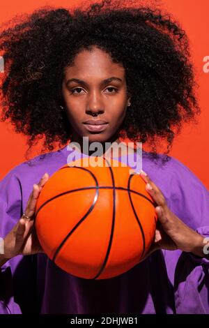 Femme ethnique sans émotion avec cheveux afro portant un maillot de sport violet et tenant un ballon de basket orange vif en studio Banque D'Images