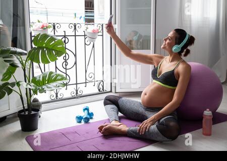 Vue latérale d'une femme enceinte souriante assise sur un tapis et prendre le selfie sur un smartphone tout en écoutant de la musique dans un casque après avoir pratiqué le yoga Banque D'Images