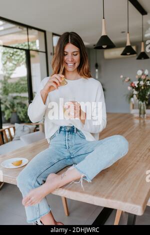 Joyeuse femelle pressant le jus de citron dans le verre avec de l'eau pendant préparation de la boisson rafraîchissante vitaminée assis sur la table à accueil Banque D'Images