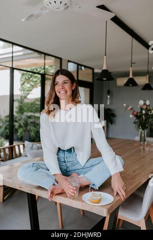 Femme joyeuse assise sur une table à la maison avec un verre de eau rafraîchissante avec jus de citron et apprécier le matin Banque D'Images