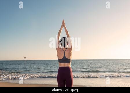 Vue arrière de la femelle non reconnaissable en casque d'activité debout dans la montagne posez avec les bras vers le haut tout en pratiquant le yoga sur une plage de sable près de la mer Banque D'Images