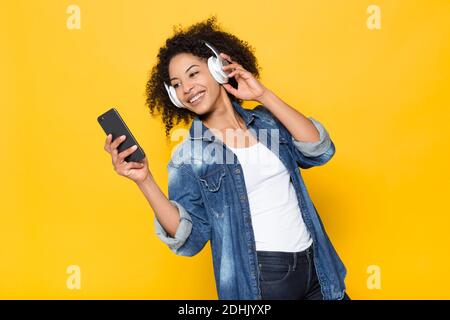 Joyeux jeune Afro-américain hipster femme en denim chemise avoir amusez-vous et profitez de votre musique à plein volume grâce à un casque sans fil sur des écouteurs lumineux fond jaune Banque D'Images