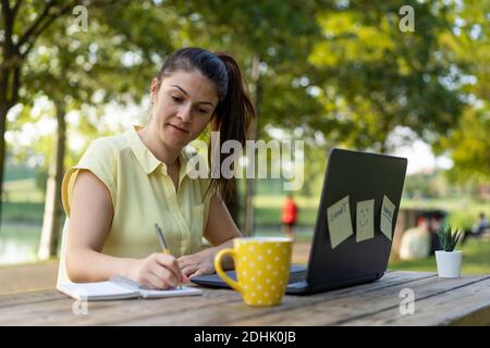 Jeune femme utilisant un ordinateur portable dans le parc public - Femme indépendante du millénaire travaillant à distance et prenant des notes dans un Parc de la ville - technologie et Banque D'Images