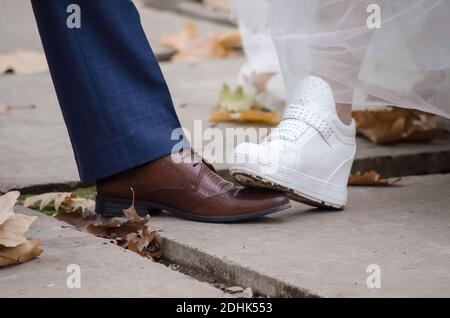 Le pied de la mariée en baskets blanches marche sur le pied du marié sur des chaussures brunes, en automne parmi les feuilles, gros plan Banque D'Images