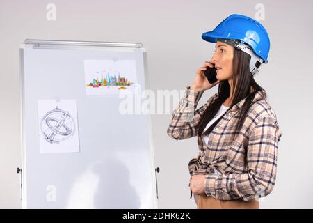 Conversation téléphonique. Femme architecte portant un casque de sécurité parlant au téléphone contre le fond des projets de travail. Banque D'Images