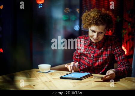 Une femme mûre fait des achats en ligne en utilisant un ordinateur tablette dans un café. Une femme plus âgée paie pour le paiement en ligne par carte de crédit. Moderne senior utilise plastique c Banque D'Images