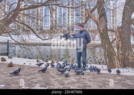 En hiver, un homme asiatique senior heureux, dans une veste en duvet, un Jean et un bonnet tricoté, nourrit les oiseaux dans un parc municipal près de la rivière. Il regarde la caméra et Banque D'Images