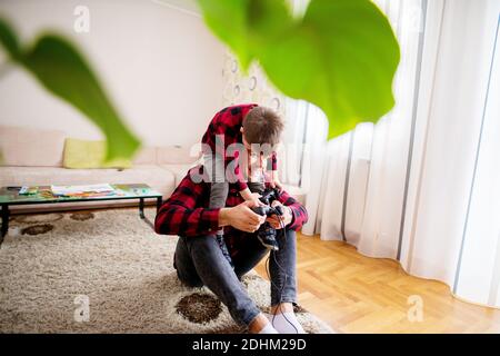 Un jeune fils joyeux se penchait sur son père tout en le tenant assis sur le sol tenant une manette dans un salon lumineux. Banque D'Images