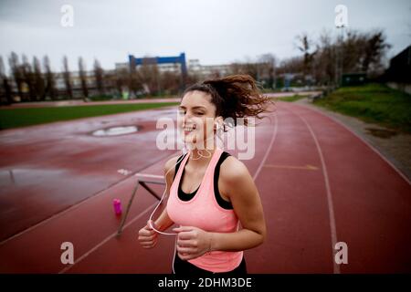 Portrait d'une jeune fille de forme physique heureuse et attrayante qui fait du jogging sur une piste tout en écoutant de la musique près du terrain de football. Banque D'Images