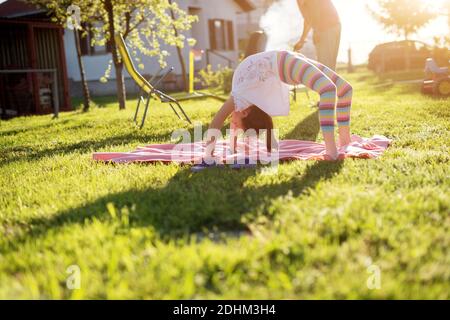 Une petite fille adorable fait un pont de yoga par une belle journée ensoleillée pendant que son père est en train de griller. Banque D'Images