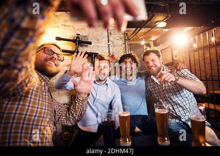 Quatre amis multiculturels souriants agitant et prenant un selfie tout en étant assis dans le pub. Banque D'Images