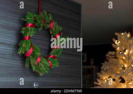 Couronne en pin foré avec ruban rouge sur l'ouverture de la porte avec arbre de noël blanc illuminé à l'intérieur de la maison. Banque D'Images