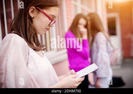 Triste fille malheureuse d'école secondaire lisant des notes d'école tandis que ses camarades la racontent devant tout en se tenant à l'extérieur de l'école. Banque D'Images