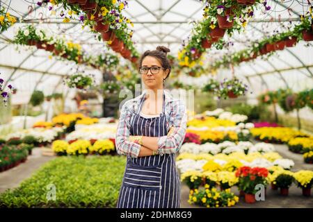 Belle charmante fleuriste femme posant avec des bras croisés dans la serre colorée et lumineuse. Banque D'Images