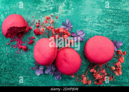 Macarons aux fraises sur fond vert intense décoré de fleurs. Composition contrastée perturbée par un biscuit en quartet avec des miettes. Banque D'Images