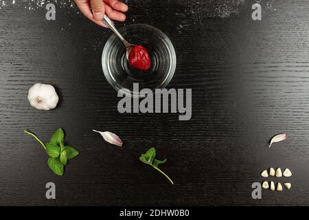 girl chef puts tomato paste in a bowl for making italian pizza at home on a black wooden kitchen table Stock Photo