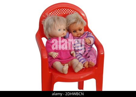 Deux vieilles poupées assises sur une chaise en plastique rouge avec un fond blanc. Une poupée fille avec une robe tricotée et un oeil endommagé. Isolé. Banque D'Images