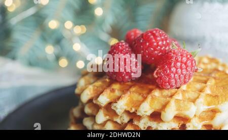 Gaufres viennoises avec framboises et miel sur la table de Noël avec branches d'arbres et guirlandes de lumières en arrière-plan. Gros plan, espace copie. Banque D'Images