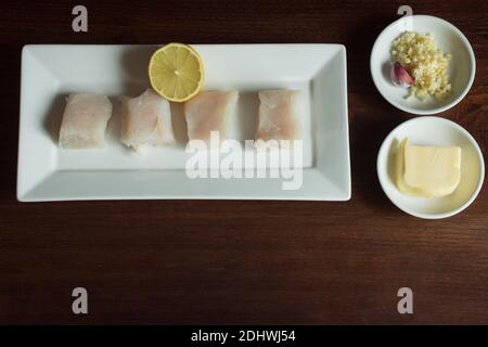 Assiette rectangulaire blanche avec morceaux de pain de merlu cru et un citron coupé en deux servi avec de l'ail haché et du beurre sur une table en bois. Recettes avec du poisson Banque D'Images