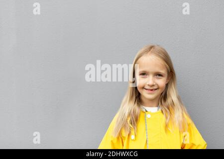 Bonne petite fille caucasienne 6-7 ans avec cheveux blonds en imperméable jaune debout à l'extérieur près du mur gris. Couleurs de l'année 2021, gris ultime Banque D'Images