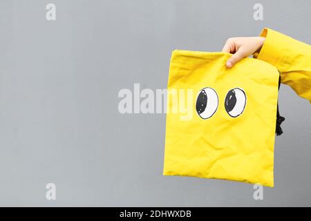 Enfant en imperméable jaune debout à l'extérieur près du mur gris et tenant un sac amusant. Couleurs de l'année 2021, gris ultime et illuminant Banque D'Images