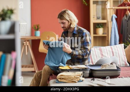 Jeune homme assis sur le canapé empaquant les vêtements dans la valise il préparation des vacances Banque D'Images