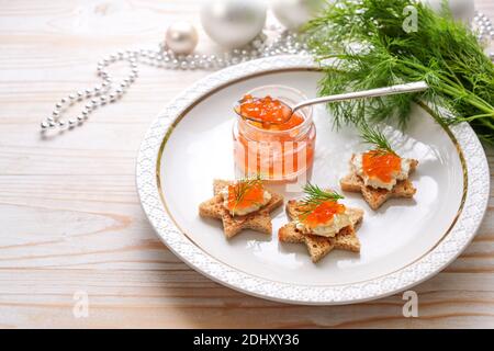 Caviar rouge à la crème et à l'aneth sur des canapés toastés en forme d'étoile sur une assiette blanche, préparation pour un buffet de fête sur une table en bois clair Banque D'Images