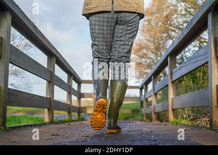 Vue arrière à angle bas de la femme britannique en bottes/bottes wellington, isolée en plein air marchant sous le soleil hivernal traversant une passerelle en bois. Banque D'Images
