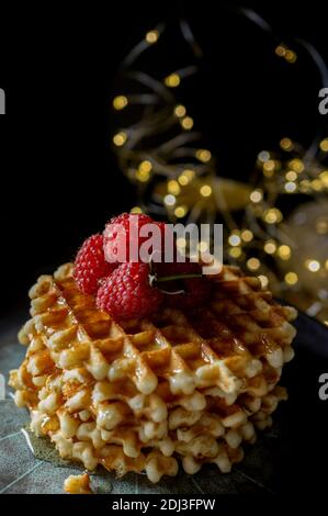 Gaufres viennoises avec framboises et miel sur la table de Noël avec branches d'arbre de Noël et guirlande lumineuse en arrière-plan. Banque D'Images