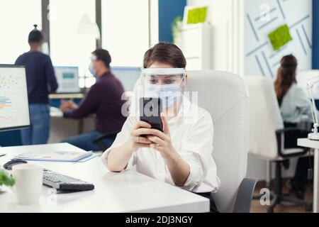 Femme d'affaires discutant sur un smartphone assis sur son lieu de travail dans un bureau occupé portant un masque facial pendant une pandémie mondiale avec covid19. Collègues multiethniques travaillant dans le respect de la distance sociale dans la société financière. Banque D'Images