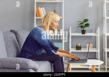 Vue latérale d'une femme mûre tapant sur un ordinateur portable tout en étant assise sur son canapé à la maison. Banque D'Images