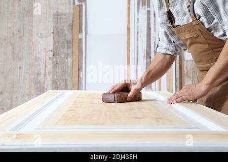 Charpentier mâle travaillant le bois dans l'atelier de menuiserie, sablant une porte en bois avec du papier de verre, portant l'ensemble Banque D'Images