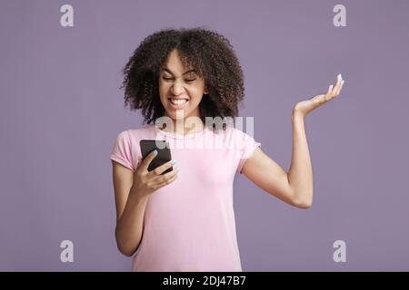 Contrarié femme afro-américaine millénaire en t-shirt rose avec des cheveux bouclés, lit le message et se met en colère Banque D'Images