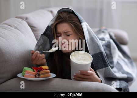 Bouleverser la jeune femme qui fait face à la dépression en mangeant de la crème glacée et des bonbons sous la couverture sur le canapé Banque D'Images