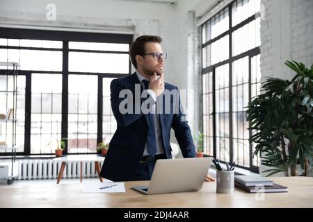 Homme d'affaires attentionné, cherchant à côté, rêvant, se tenant près du bureau Banque D'Images