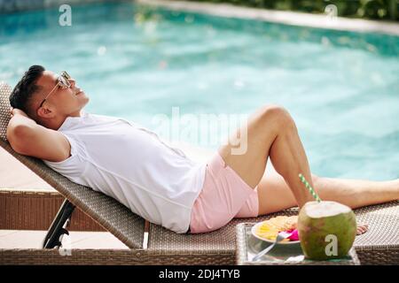 Joyeux jeune homme en lunettes de soleil se reposant sur une chaise longue en nageant piscine avec cocktail de noix de coco et assiette de fruits sur la table à proximité Banque D'Images