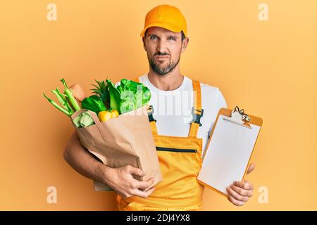 Jeune homme beau portant un uniforme de messagerie avec des articles d'épicerie et un presse-papiers expression sans indice et confuse. Notion de doute. Banque D'Images