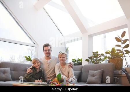 Portrait grand angle d'un père aimant embrassant deux fils et regardant l'appareil photo tout en étant assis sur un canapé dans un intérieur moderne, espace de copie Banque D'Images