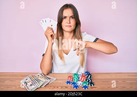 Belle femme caucasienne jouant au poker tenant des cartes avec le visage en colère, signe négatif montrant l'aversion avec les pouces vers le bas, concept de rejet Banque D'Images