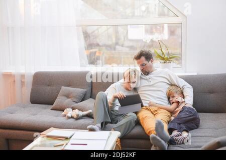 Portrait grand angle de père moderne mature allongé sur un canapé avec deux fils et regardant des vidéos ensemble via une tablette, espace de copie Banque D'Images